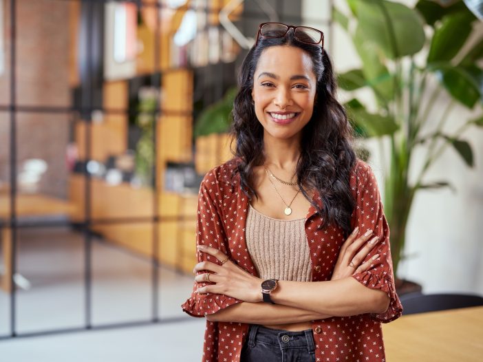 Portrait of smiling young multiethnic woman looking at camera with crossed arms. Successful latin business woman standing in modern office with copy space. Young university hispanic girl with pleasant smile and folded arms.